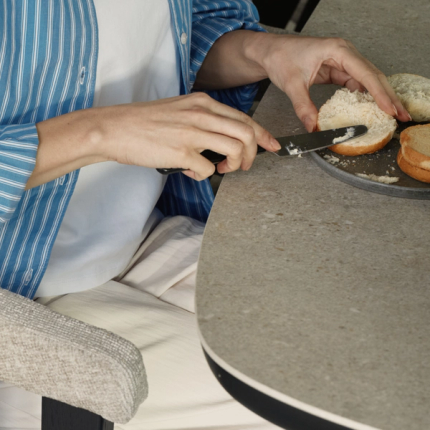 Persoon aan tafel die een broodje besmeert, met gestoffeerde stoel en afgerond tafelblad in beeld.