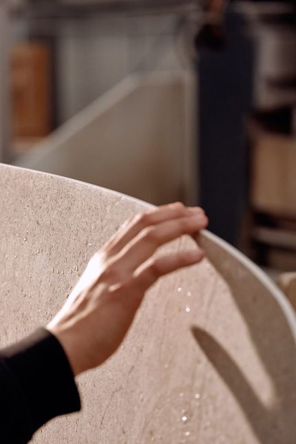 Hand van persoon die het afgeronde blad van een lichte keramische tafel aanraakt, met zachte lichtinval op het oppervlak.