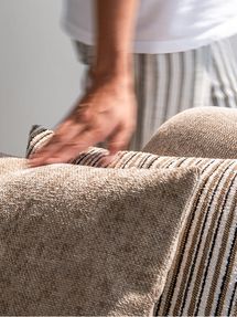 Gros plan d’une main glissant sur un canapé beige à la riche texture tissée, agrémenté de coussins décoratifs rayés.