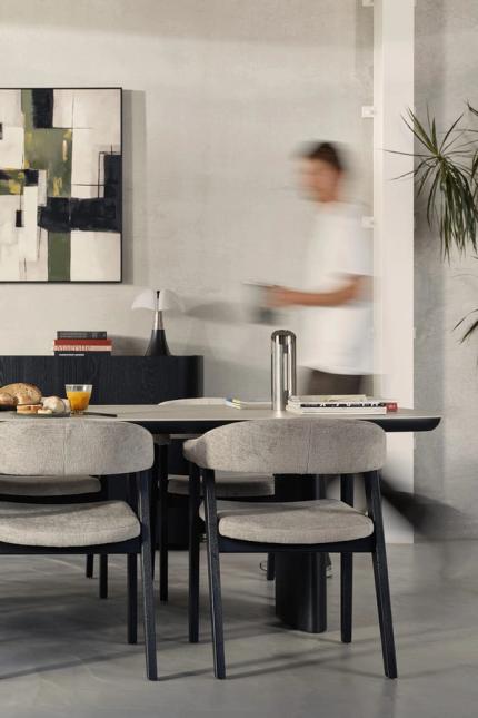 Table à manger avec chaises rembourrées grises dans un intérieur minimaliste ; petit-déjeuner sur la table et une personne passe en flou.