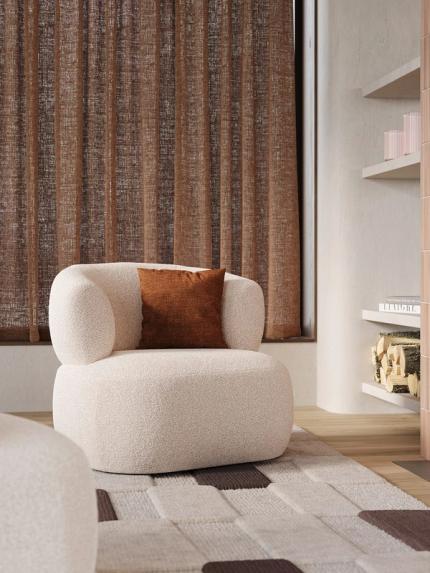 Fauteuil Bomba en tissu bouclé clair avec coussin brun rouille, sur un tapis texturé devant des rideaux bruns dans un coin salon.