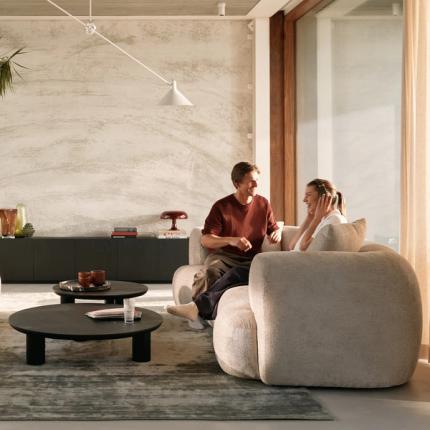 Zachte, ronde zetel in lichte stof met twee lage, ronde zwarte salontafels in een warme, minimalistische woonkamer.