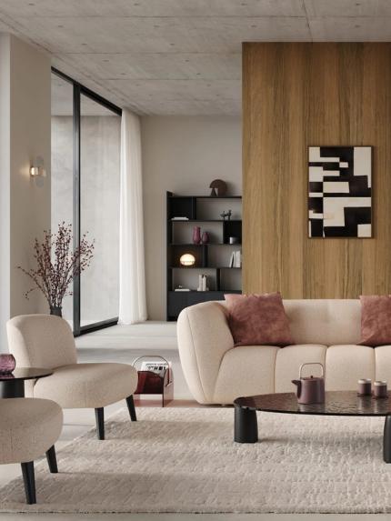 Fauteuil Aviano en tissu clair avec pieds noirs, dans un salon contemporain avec canapé beige, table basse noire et cheminée.