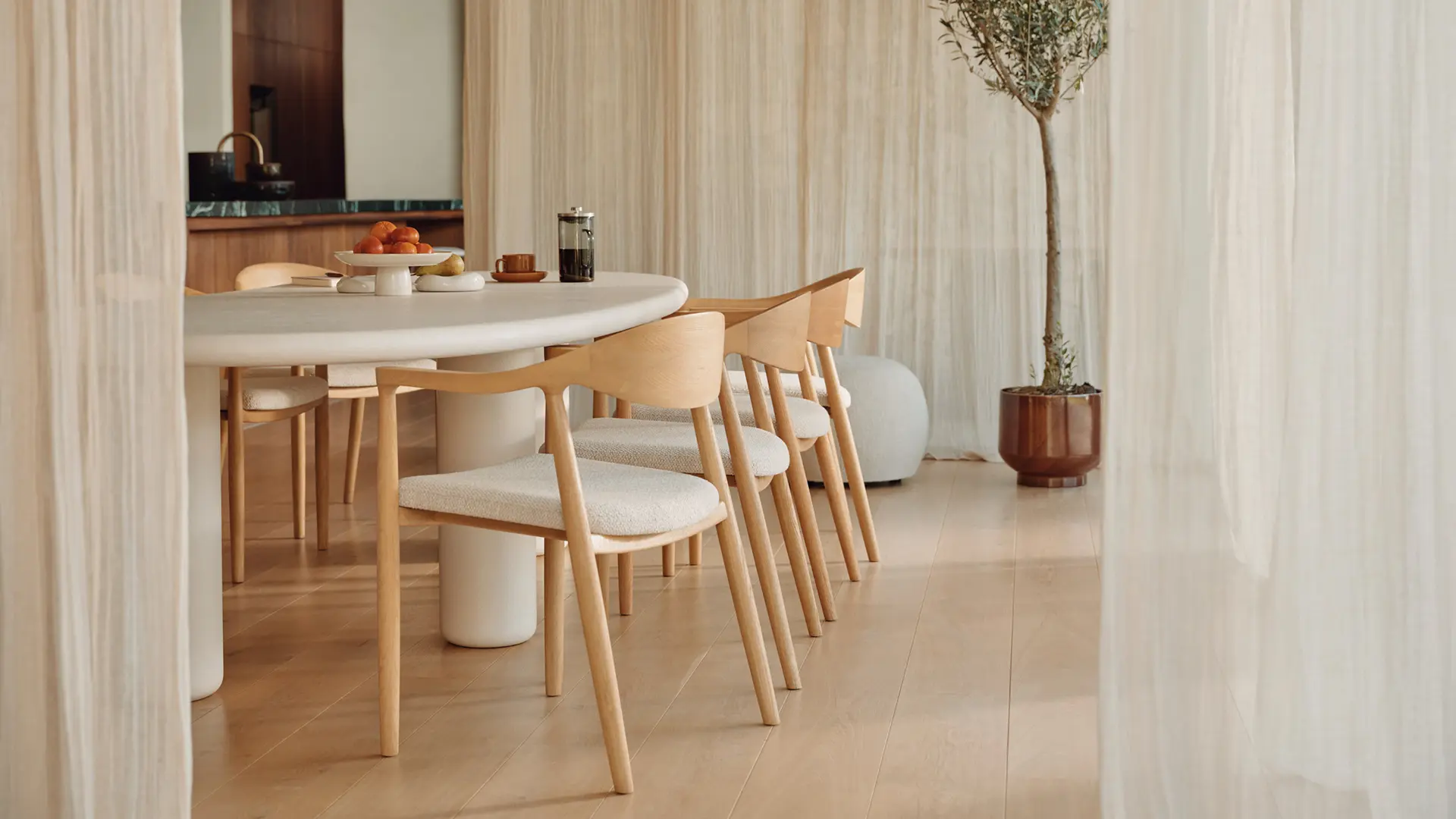 Chaises en bois clair avec assise beige autour d’une table ovale blanche, derrière des voilages avec un arbre en pot.