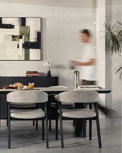 Twee licht gestoffeerde eetkamerstoelen met afgeronde rugleuning en donkerhouten poten staan aan een moderne eettafel, terwijl een persoon op de achtergrond voorbijloopt.