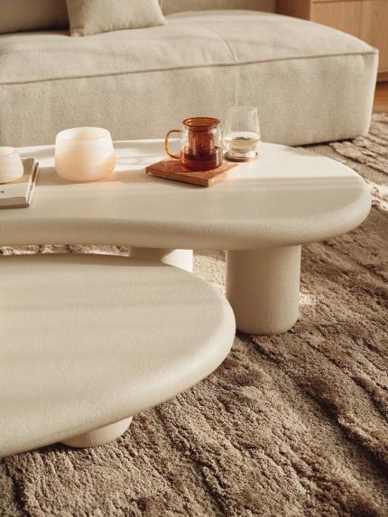 Deux tables basses aux formes organiques, finition claire, sur un tapis texturé, avec verres, bougie et plateau en bois.