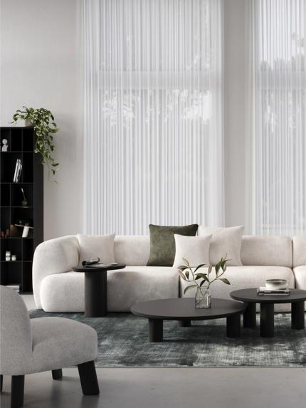 Canapé d'angle clair avec des coussins blancs et vert olive, lampe arc et tables basses rondes noires sur un tapis bleu-vert.