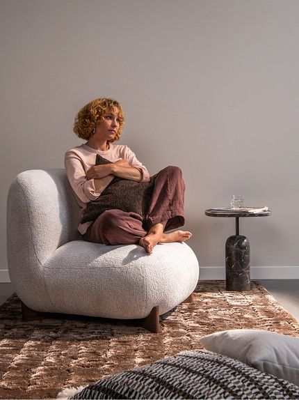 Personne assise en tailleur dans un fauteuil arrondi de couleur claire, tenant un coussin foncé, à côté d’une table d’appoint en marbre sur un tapis brun moelleux.