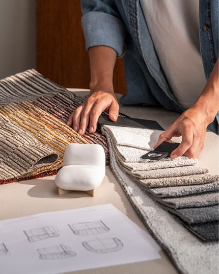 Deux personnes comparent des échantillons de tissus d’ameublement de différentes couleurs et textures, à côté de croquis de design et d’échantillons de matériaux sur une table.