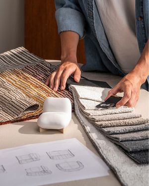 Deux personnes comparent des échantillons de tissus d’ameublement de différentes couleurs et textures, à côté de croquis de design et d’échantillons de matériaux sur une table.