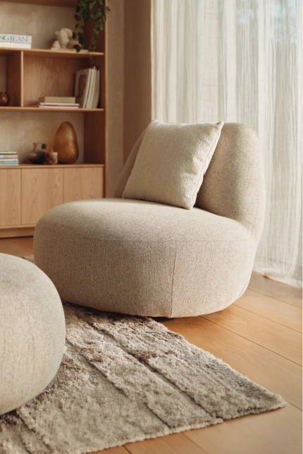 Beige bouclé fauteuil met kussen en bijpassende poef in rustige woonkamer met houten kast en zacht daglicht