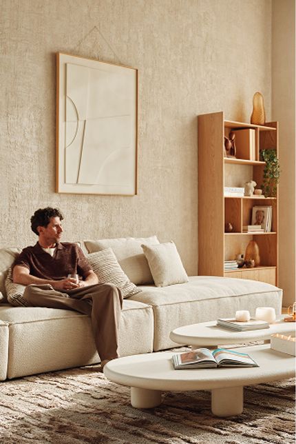 Man op lichte zetel in woonkamer met houten boekenkast, witte salontafels en groot raam.