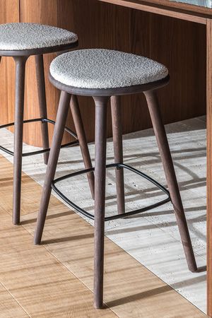 Deux tabourets de bar hauts avec pieds en bois foncé, repose-pieds en métal et assises claires rembourrées au revêtement doux et texturé.