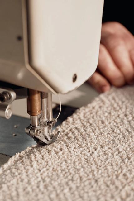 Close-up van een naaimachine die een dikke, beige bouclé stof stikt; handen begeleiden de stof langs de naald.