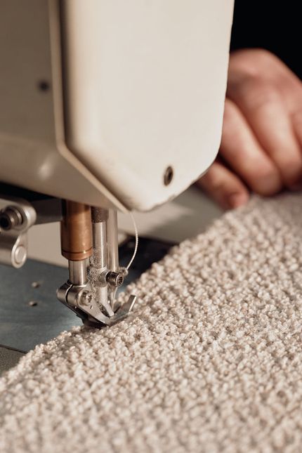 Close-up van een naaimachine die een dikke, beige bouclé stof stikt; handen begeleiden de stof langs de naald.