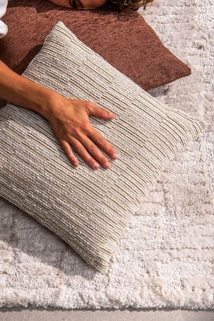Gros plan d’une main posée sur un coussin clair aux fines rayures et à la riche texture tissée, à côté d’un coussin brun rouille sur un tapis doux.