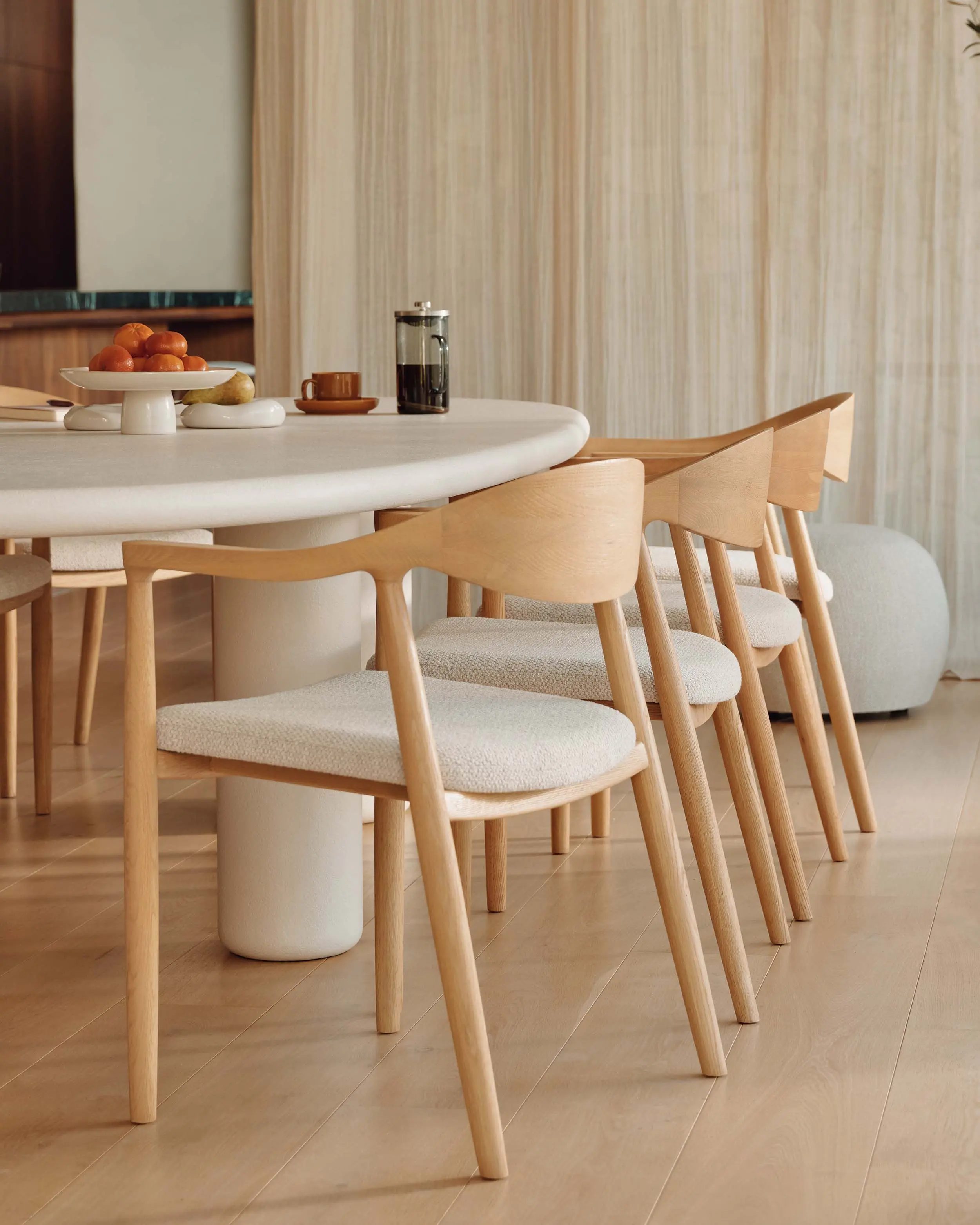 Eethoek met ronde tafel op centrale poot, houten stoelen met stoffen zitting en lichte gordijnen in warme keukenruimte.