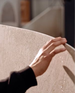 Gros plan d’une main glissant sur le bord arrondi d’un plateau de table clair, à l’aspect minéral et à la texture subtile.