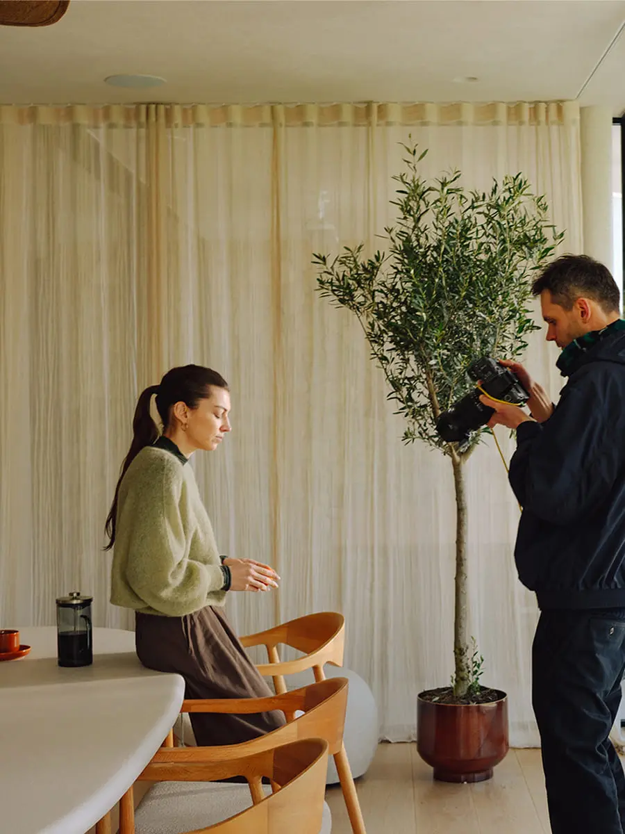 Fotograaf maakt beelden van een vrouw aan de eettafel in een lichte ruimte met houten stoelen, fruit en kamerplant bij het raam.