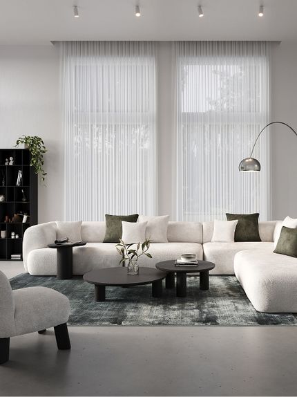 Salon avec canapé modulaire Tortona en tissu gris clair, tables basses rondes noires, tapis vert et grandes fenêtres avec rideaux légers.