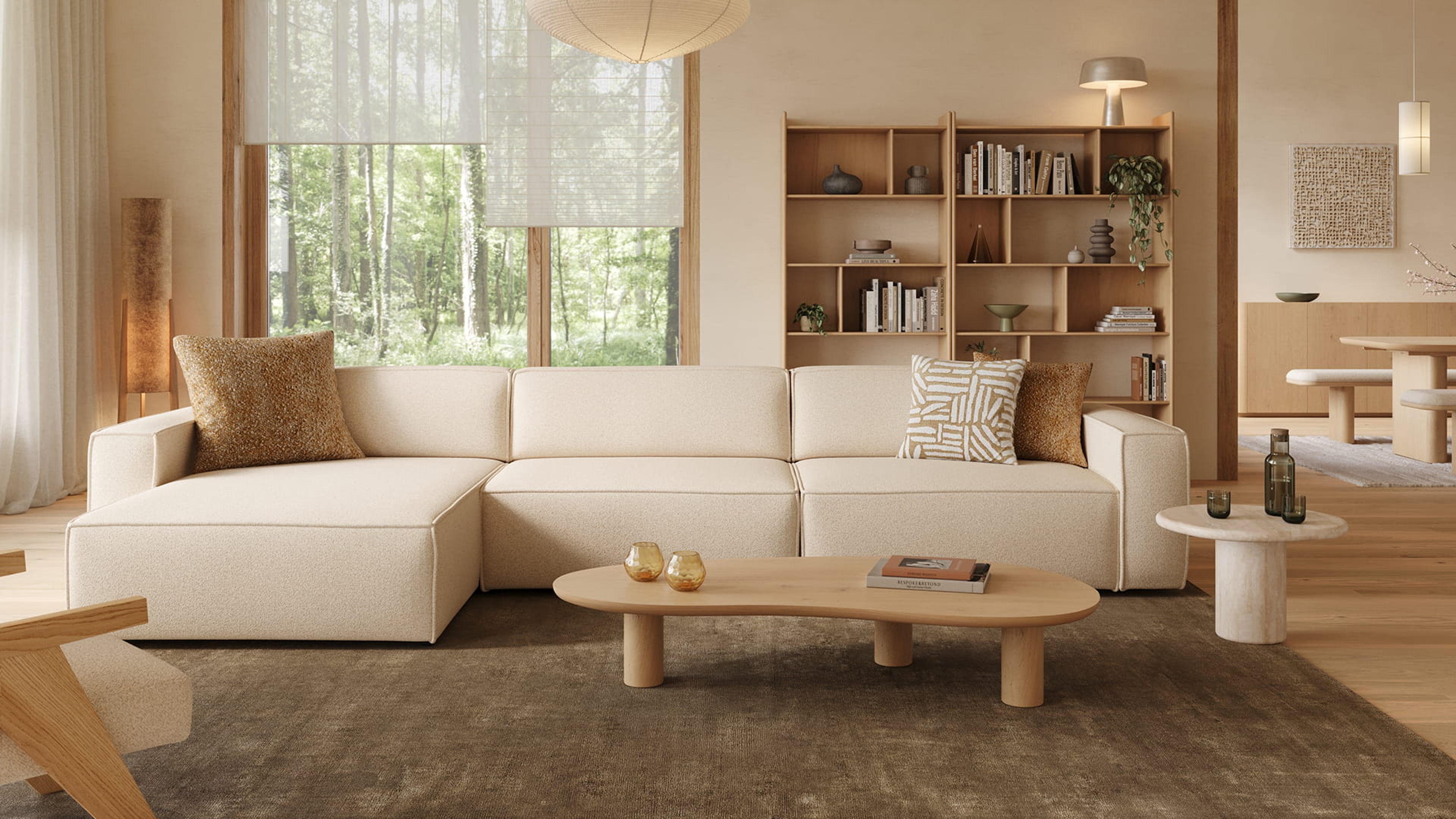 Canapé d’angle Isola beige dans un salon lumineux avec grandes fenêtres, bibliothèque en bois, table basse et matériaux naturels.