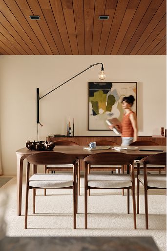Houten eettafel met zes stoelen, boeket en booglamp; persoon loopt voorbij in lichte eetkamer.