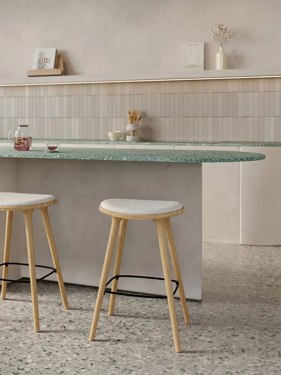 Afgerond keukeneiland met groen gespikkeld werkblad en verticale wandtegels, met twee houten barkrukken met licht gestoffeerde zitting op een terrazzo vloer.