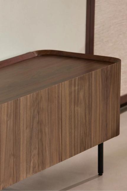 Close-up van een houten dressoir met geribbelde fronten en afgeronde rand, op slanke zwarte pootjes tegen een rustige wand.