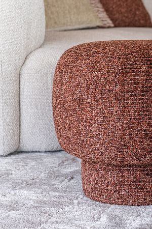 Gros plan d’un pouf arrondi en tissu brun rouille richement tissé, placé devant un canapé clair sur un tapis doux.