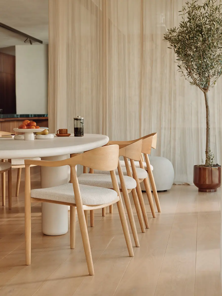 Chaises en bois clair avec assise beige autour d’une table ovale blanche, derrière des voilages avec un arbre en pot.