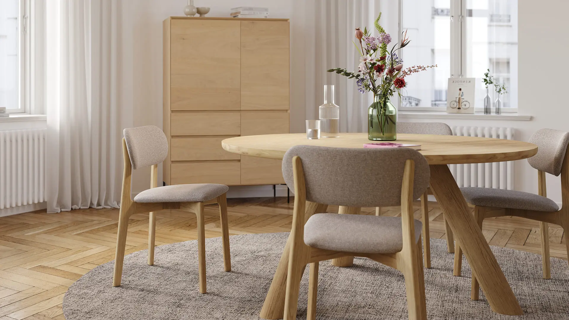 Salle à manger lumineuse avec table ronde en bois et chaises rembourrées, un vase en verre vert avec fleurs sur la table et un meuble en bois à l’arrière-plan.
