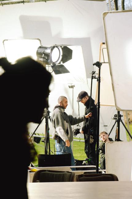 Silhouette d’une personne au premier plan, avec une équipe de tournage et de grands panneaux lumineux à l’extérieur sur un plateau en pleine verdure.