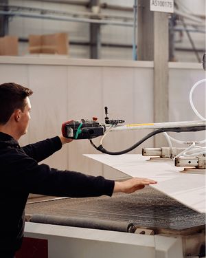 Medewerker bedient een machine in een meubelatelier en begeleidt een lichtgekleurd paneel tijdens het productieproces.