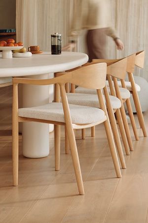 Salle à manger lumineuse avec des chaises en bois aux dossiers arrondis et aux assises rembourrées claires autour d’une table ovale, tandis qu’une personne passe à l’arrière-plan.