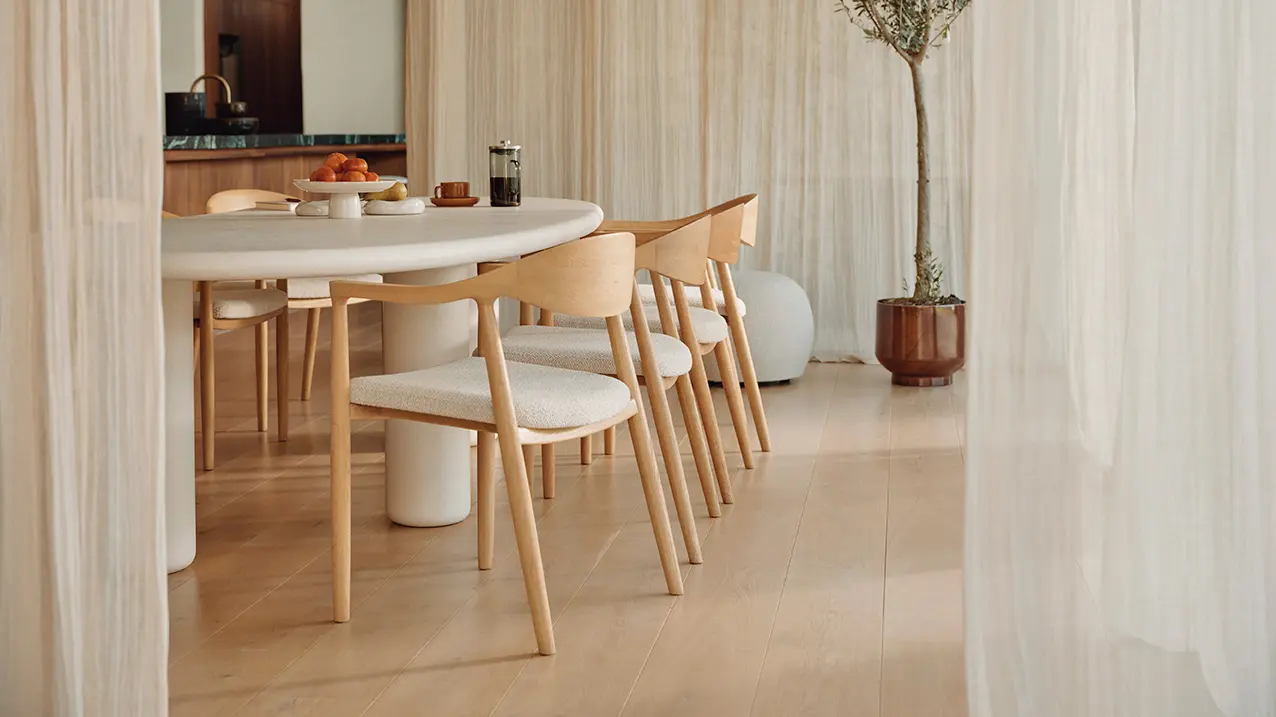 Licht houten stoelen met beige zitting rond een ovale witte eettafel, achter transparante gordijnen met kamerboom.