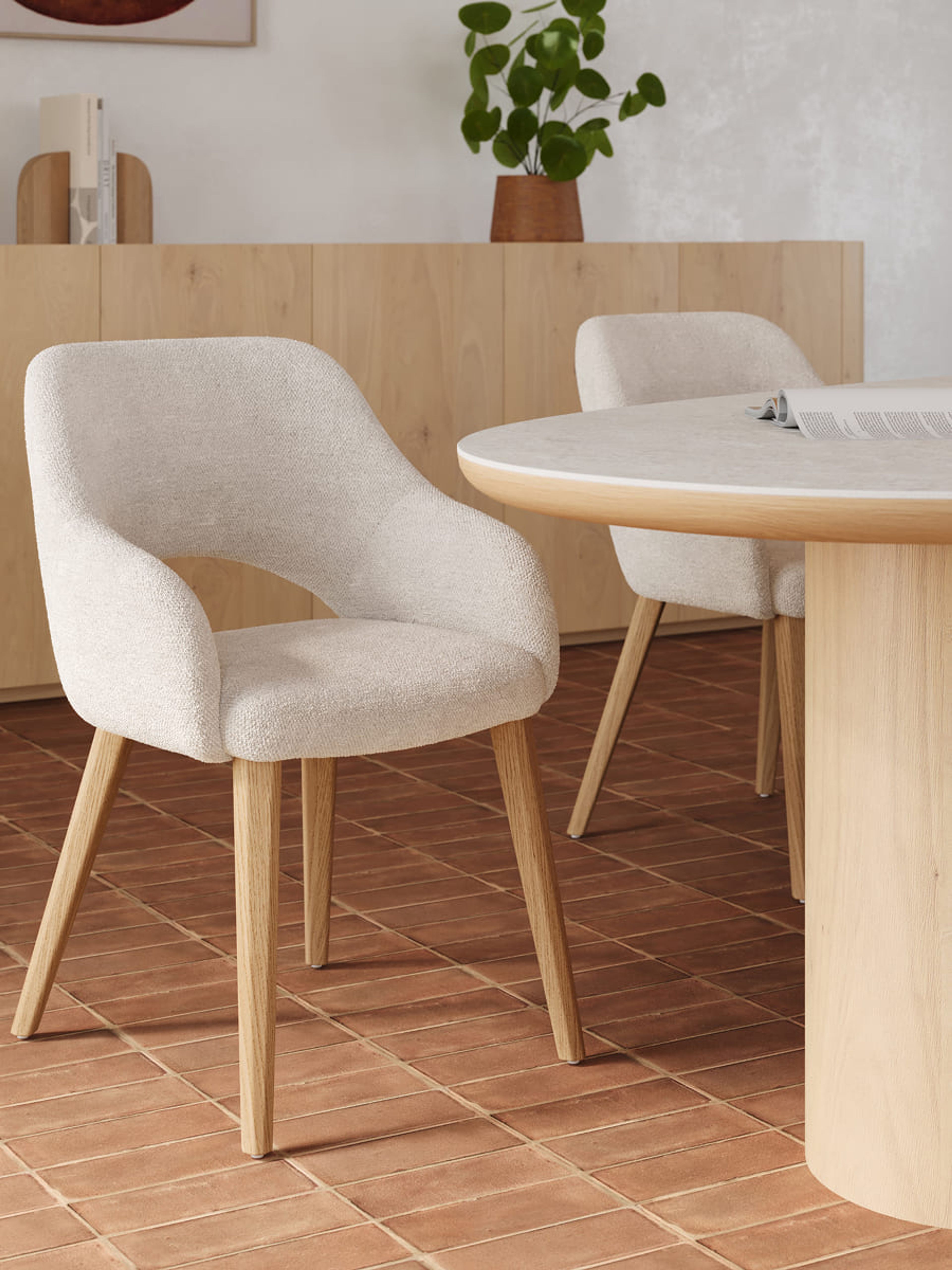 Licht gestoffeerde eetkamerstoel met houten poten naast een ronde tafel in licht hout, in een warme eetruimte met houten dressoir.