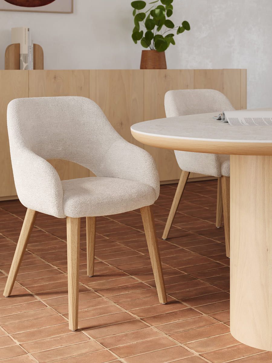 Licht gestoffeerde eetkamerstoel met houten poten naast een ronde tafel in licht hout, in een warme eetruimte met houten dressoir.
