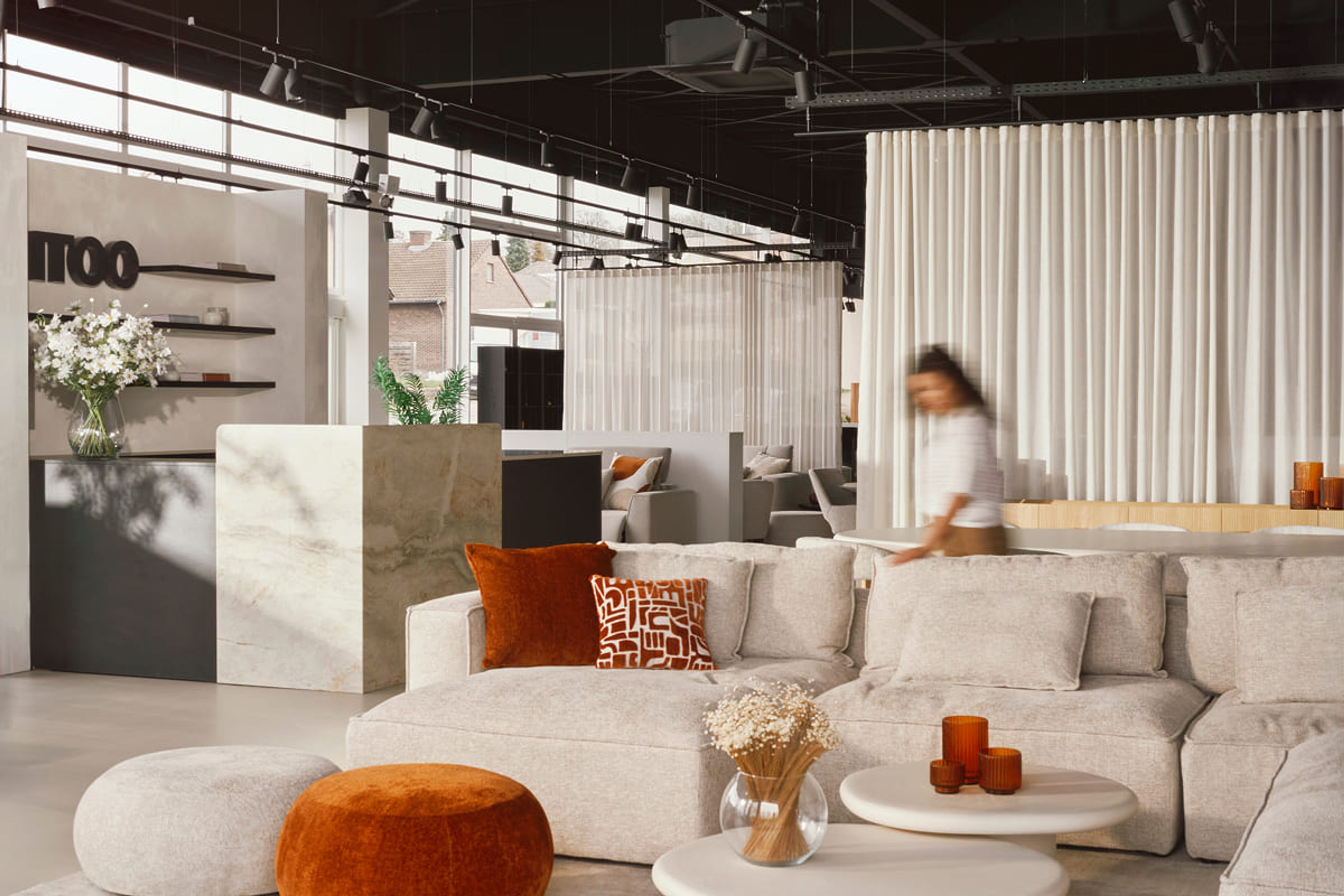 Showroom met lichte banken, oranjerode kussens en decoratieve objecten, met een persoon die door de ruimte beweegt, achter een marmeren balie.