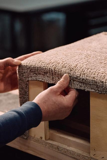 Handen trekken meubelstof strak rond een houten frame van een zitmeubel tijdens het stofferen.