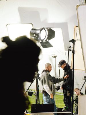 Silhouette d’une personne au premier plan, avec une équipe de tournage et de grands panneaux lumineux sur un set en extérieur, au vert.