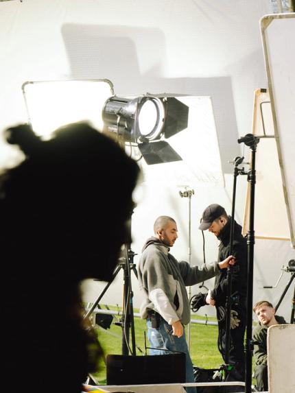 Silhouette d’une personne au premier plan, avec une équipe de tournage et de grands panneaux lumineux sur un set en extérieur, au vert.