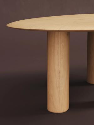 Close-up van een licht houten eettafel met afgerond tafelblad en cilindervormige poten tegen een donkerbruine achtergrond.