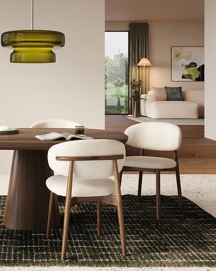Salle à manger avec table ronde en bois, quatre chaises claires et suspension verte, sur tapis foncé près d’un meuble en bois et d’une étagère ouverte.