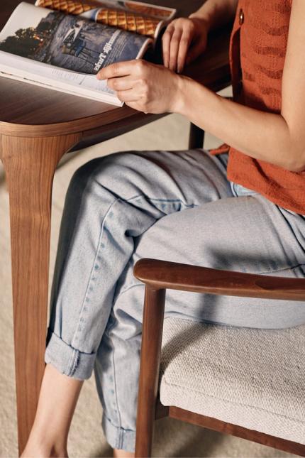 Une personne lit un magazine ouvert à une table en bois, assise sur une chaise avec accoudoirs en bois et une assise en tissu clair.