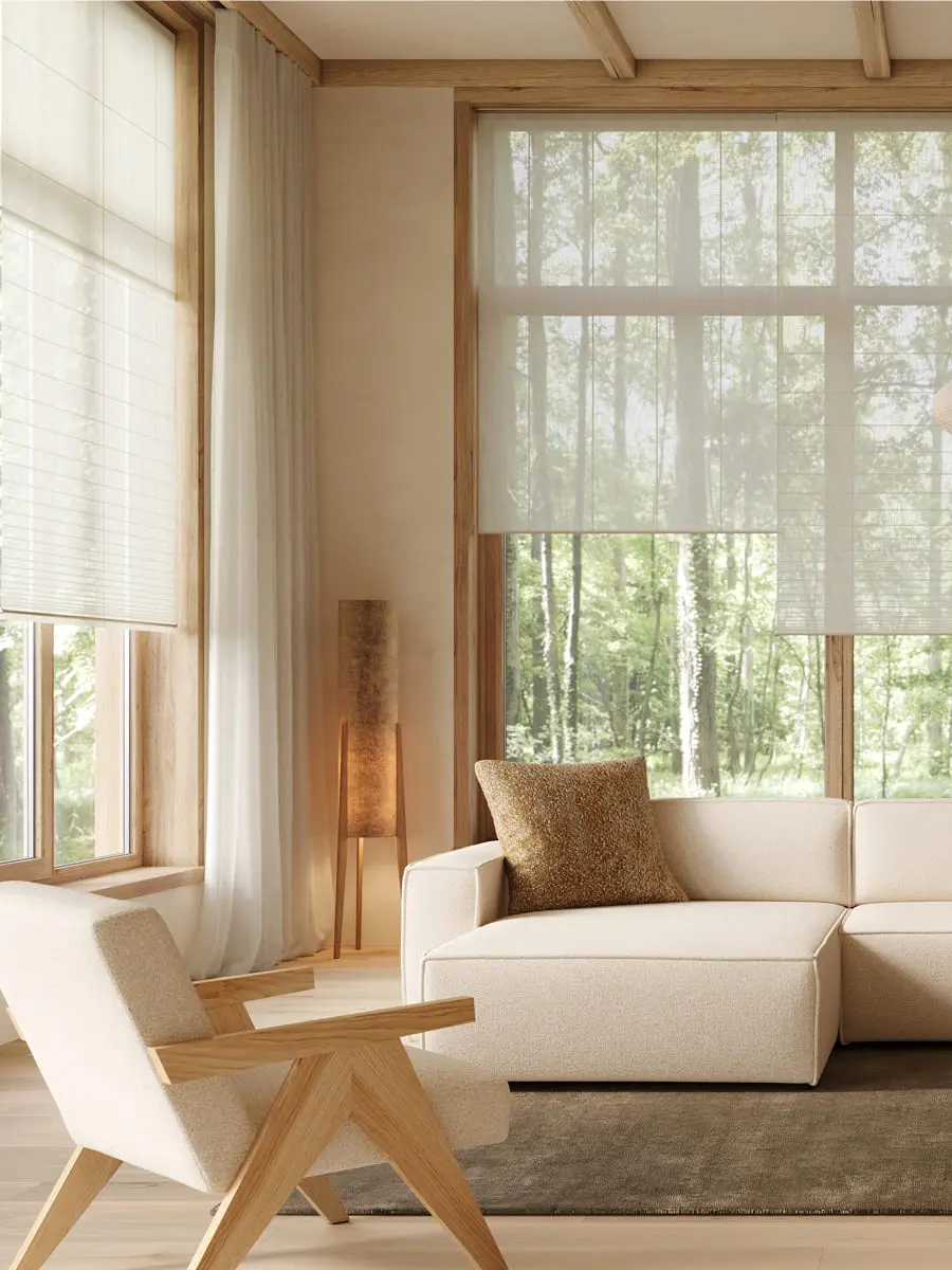 Coin salon lumineux avec un canapé beige et un fauteuil en bois sur un tapis foncé, près de grandes fenêtres donnant sur une forêt verdoyante et de voilages légers.