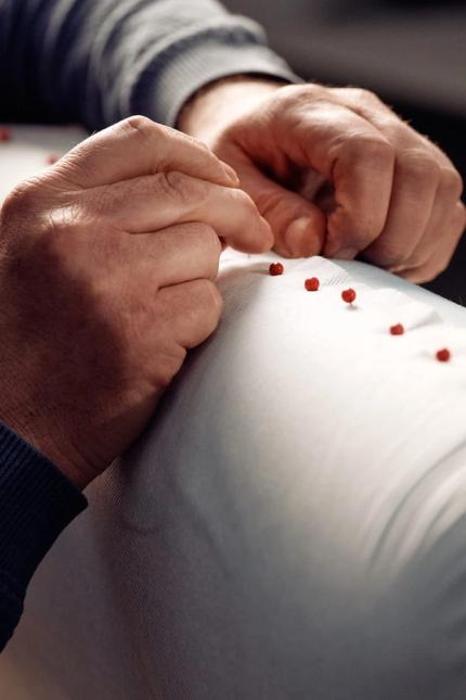Mains d'une personne piquant des épingles rouges dans un tissu clair, gros plan sur le détail du revêtement.