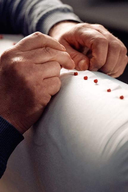 Mains d'une personne piquant des épingles rouges dans un tissu clair, gros plan sur le détail du revêtement.