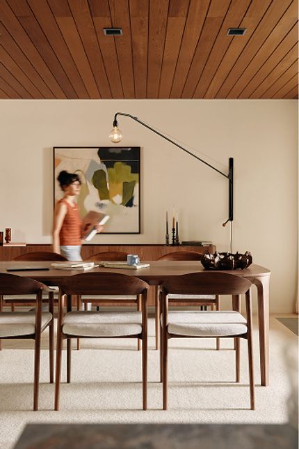 Eetkamer met lange houten eettafel en stoelen met lichte zittingen, met bloemenvaas en servies; persoon loopt voorbij bij het dressoir.