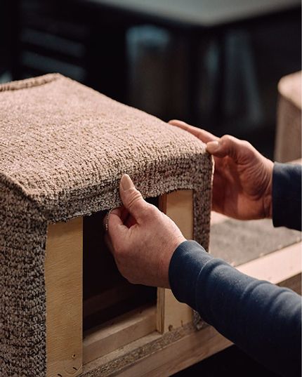 Handen die meubelstof strak over een houten frame trekken tijdens het stofferen in het atelier.