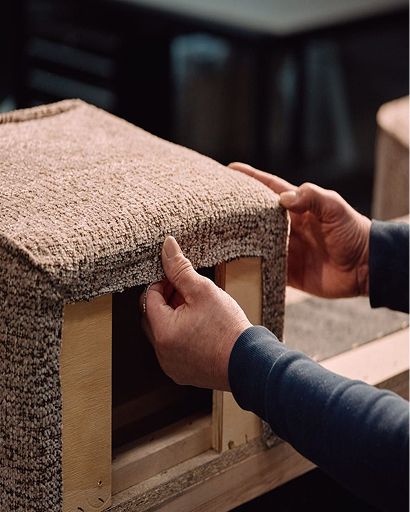 Handen die meubelstof strak over een houten frame trekken tijdens het stofferen in het atelier.